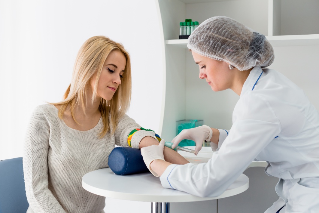 Blood test from vein with beautiful young blond woman by female doctor in white coat medical uniform on the table in white bright room.
